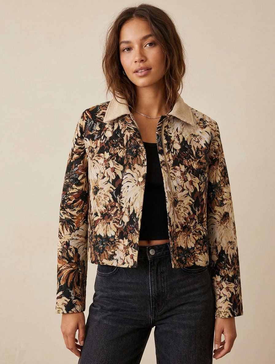 A woman modeling a cropped floral jacket with a beige collar, featuring a black, beige, and brown botanical pattern, paired with a black top and dark jeans against a solid beige background.